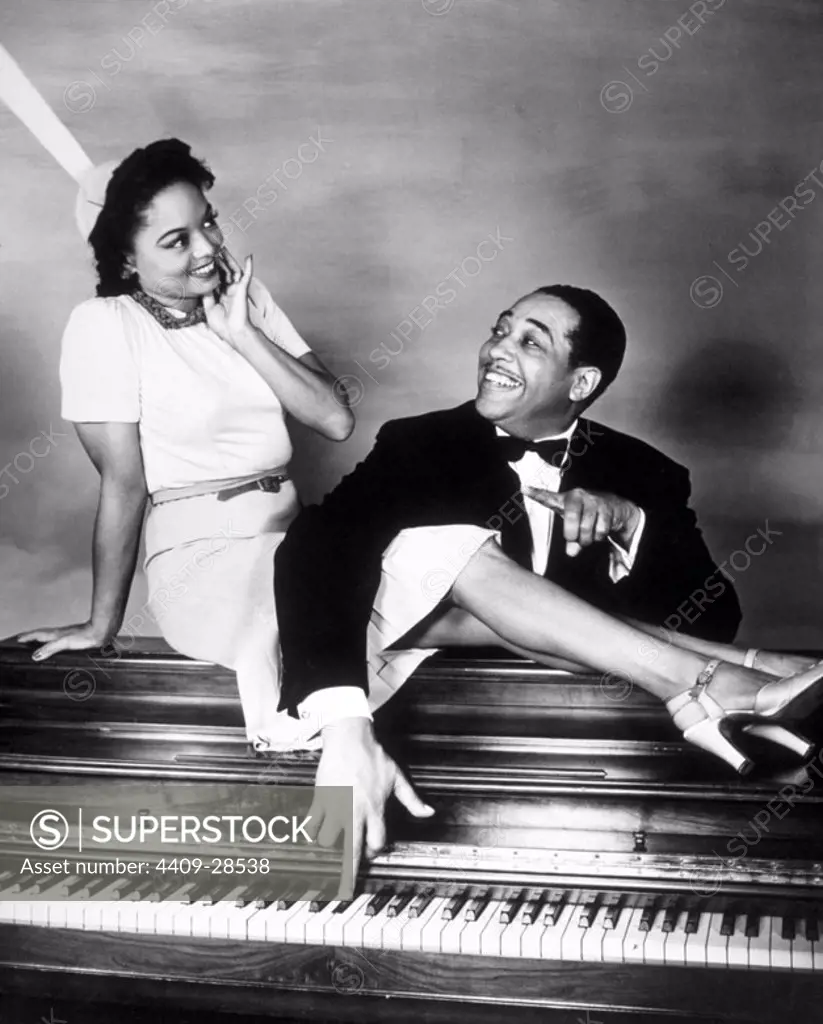 Portrait of American jazz musician Duke Ellington standing behind an upright piando and reachin over the logs of actress Louise Franklin, sitting a top the piano, to play an E Flat. Los Angeles, California, 1941.