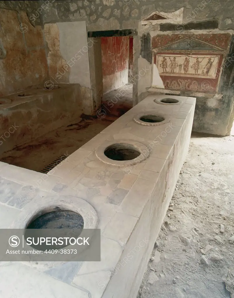Thermopolium of Asellina, tavern specializing in the sale of hot beverages. Stone bar with holes to place the amphorae containing drinks. At the bottom, fresco depicting Genius of the Pater Familias between lares. Pompeii. Italy.