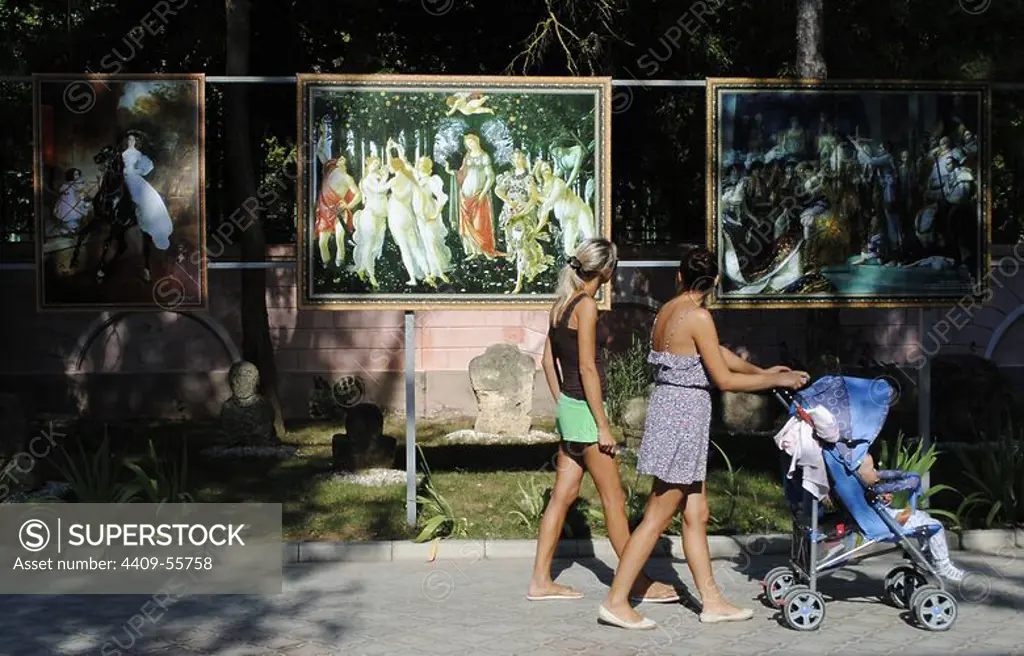 Ukraine. Autonomous Republic of Crimea. Yevpatoria. Women with a baby stroller walking down a street next to an exhibition of copies of famous paintings.
