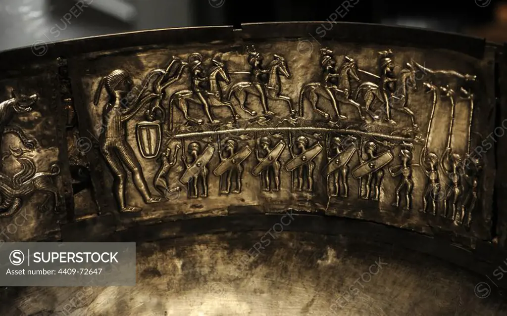 The Gundestrup cauldron. Decorated silver vessel, thought to date between 200 BC and 300 AD, placing it within the late La Te_ne period or early Roman Iron Age. Gundestrup in the Aars parish of Himmerland, Denmark. Warrior's initiation. Bottom: row of warriors bearing shields supporting a tree on their spears accompanied by three figures playing boar-headed carnyxes. Above: the tree ride four wariors, each with a boar-crested helmet. On the left-hand is a giant figure who is immersing a figure in a cauldron. National Museum of Denmark.