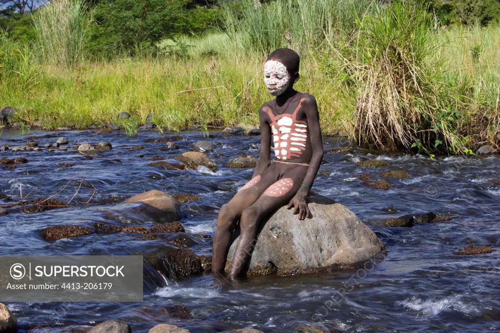 Body-painted Surma boy sitting on a rock in a river Ethiopia