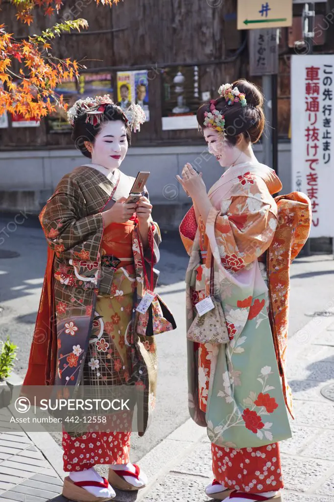 Two Geisha women dressed in kimono using mobile phones, Higashiyama Ward, Kyoto Prefecture, Kinki Region, Honshu, Japan