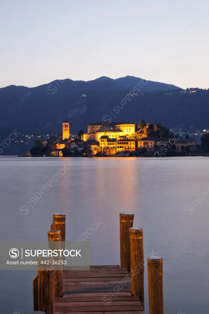 Pier in a lake with town on an island lit up at dusk, San Giulio Island, Lake Orta, Piedmont, Italy