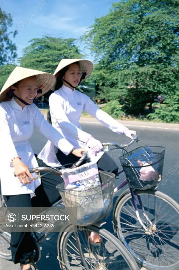 Side profile of two teenage girls wearing traditional conical hats cycling, Vietnam