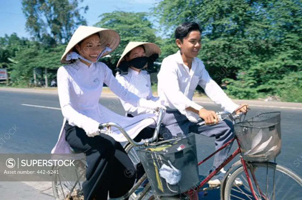 Side profile of two teenage girls wearing traditional conical hats and a teenage boy cycling, Vietnam