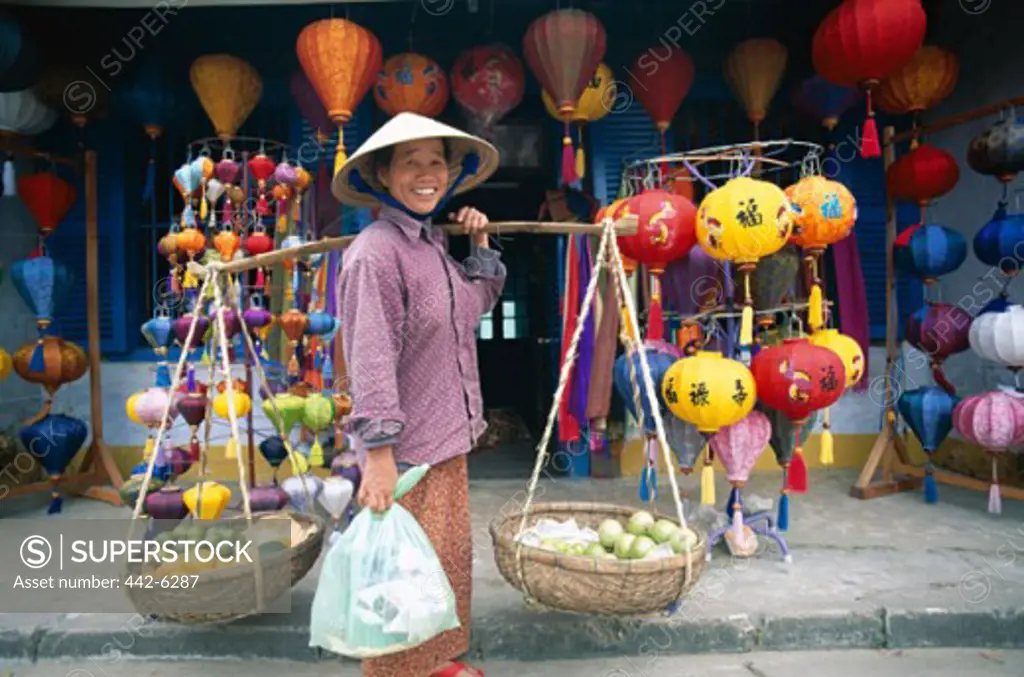 Mid adult woman carrying baskets of fruit on her shoulders standing in front of a lantern shop, Hoi An, Vietnam