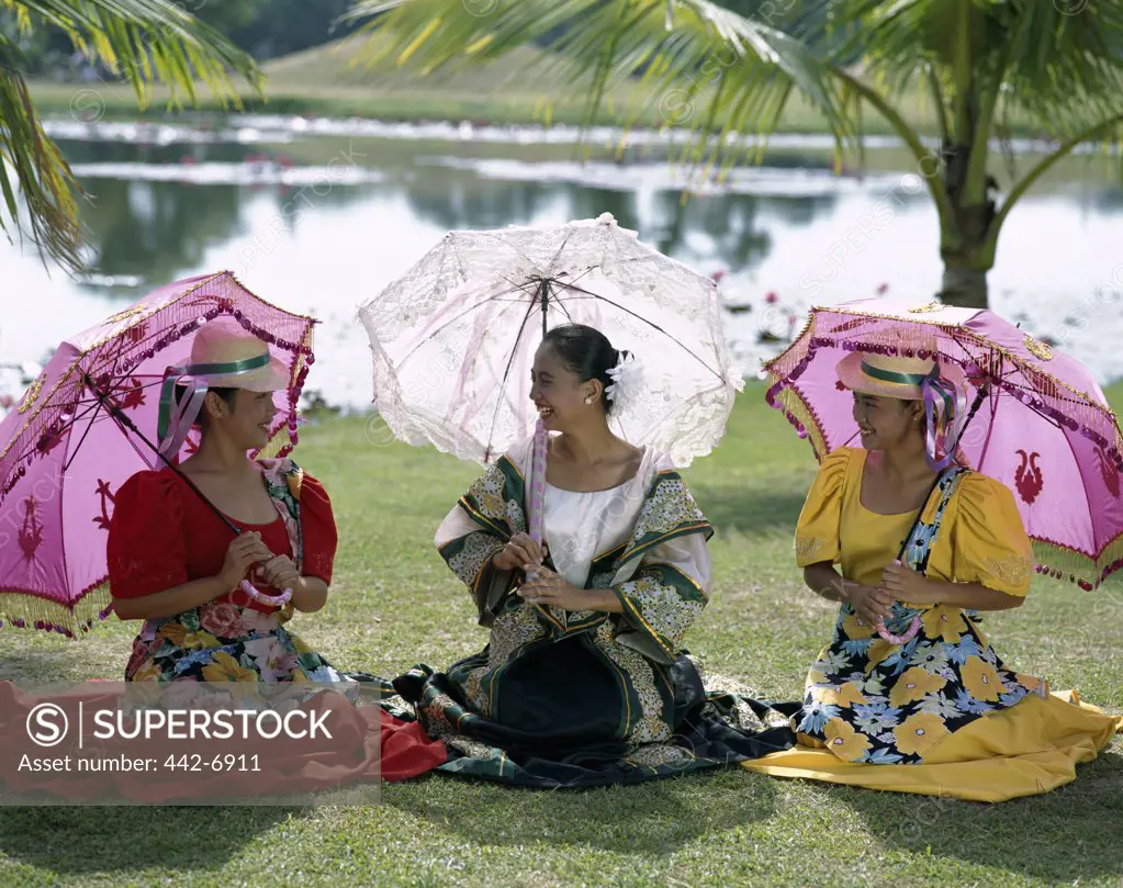 Three young women dressed in traditional costumes sitting together on a lawn, Manila, Philippines