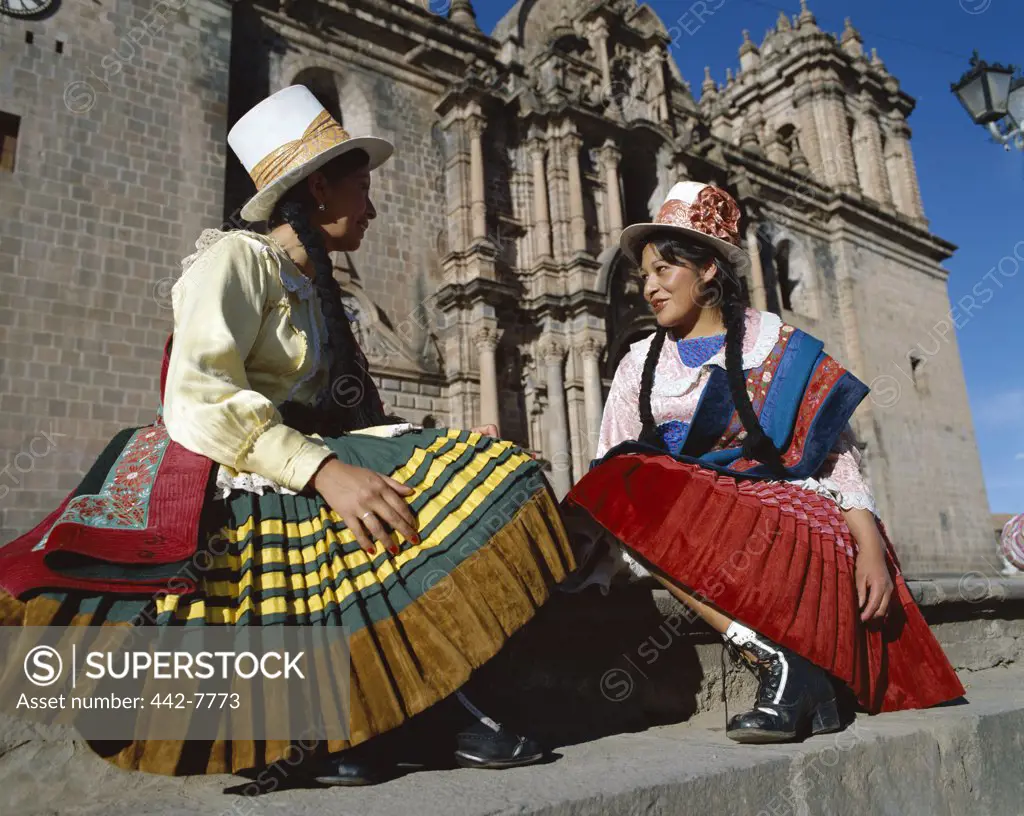 Two mid adult women dressed in traditional costumes sitting in front of a church, Cuzco, Peru
