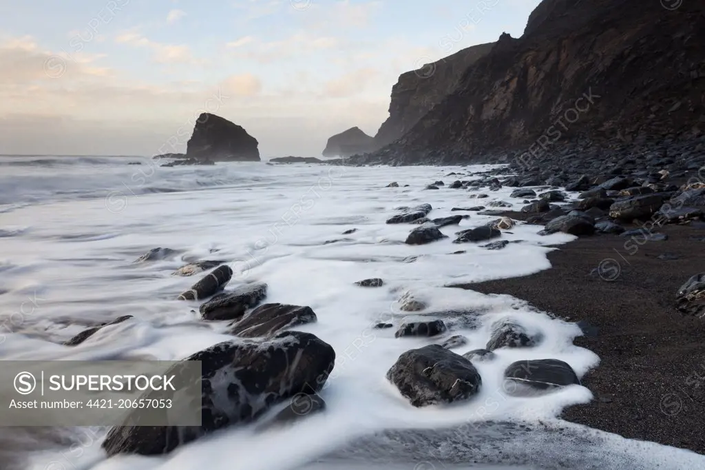 View of sea cliffs and beach with tide washing back over rocky black shingle beach on windy evening, The Strangles, North Cornwall, England, March