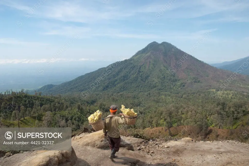 Local man carrying blocks of sulphur in baskets down from crater, with volcano in background, Mount Ijen, East Java, Indonesia