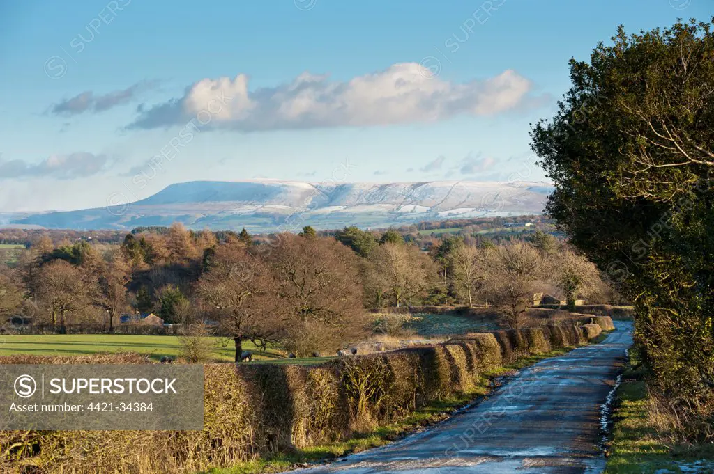 View of icy rural road, hedgerow and bare trees towards snow covered hill in distance, looking from Little Bowland Road toward Pendle Hill, Chipping, Lancashire, England, december
