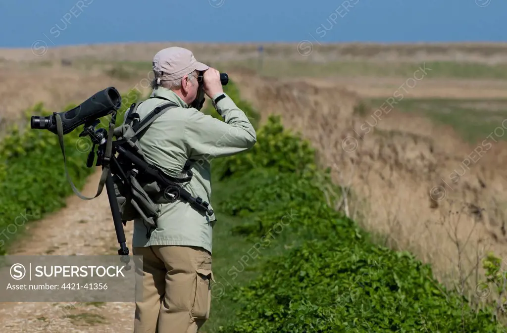 Mature birdwatcher with binoculars and telescope, looking over coastal marshland, Cley Marshes Reserve, Cley-next-the-sea, Norfolk, England, april