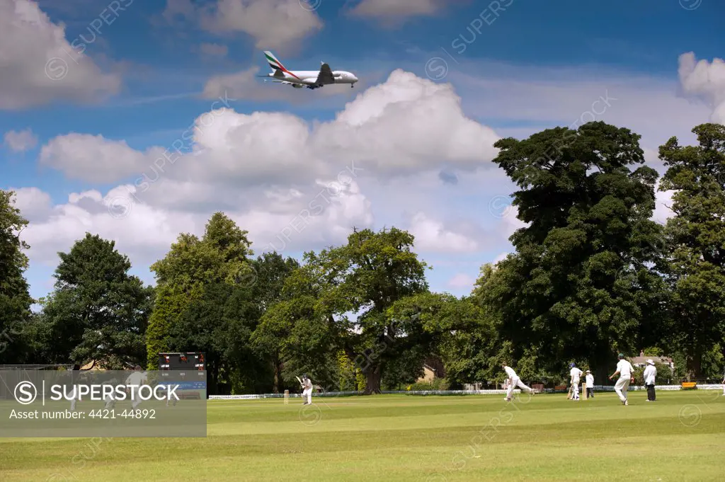 Cricket match being played on village pitch, on summer afternoon, underneath flight path to Manchester International Airport, England, July