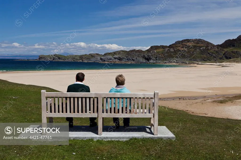 Couple sitting on bench overlooking coastline with sandy beach, Kiloran Bay, Isle of Colonsay, Inner Hebrides, Scotland