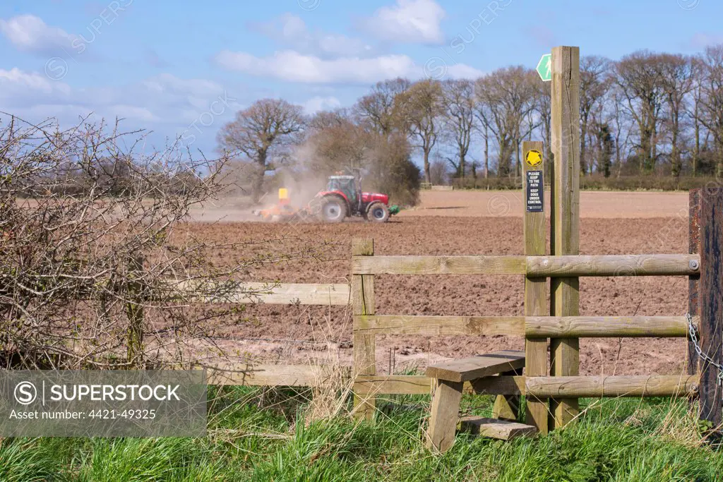 Public footpath sign and stile at edge of field, with Massey Ferguson tractor cultivating and drilling grass seed during dry dusty conditions in background, Oulton, Cheshire, England, April