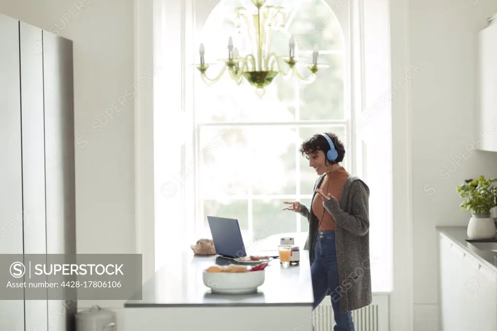 Woman with headphones video conferencing at laptop in kitchen