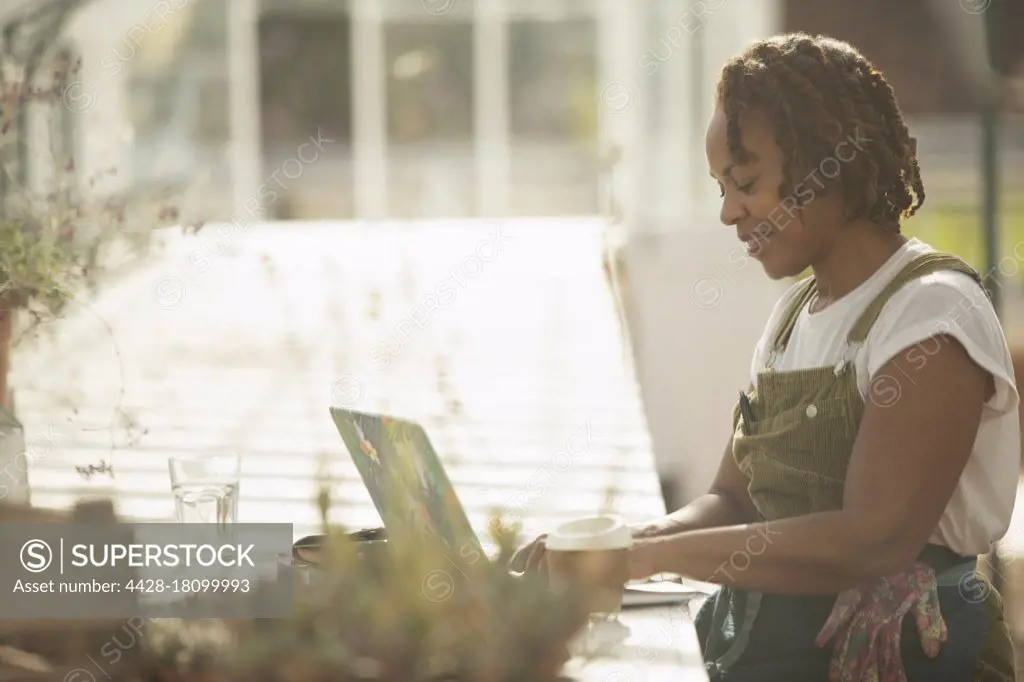 Female garden center owner working at laptop in sunny greenhouse