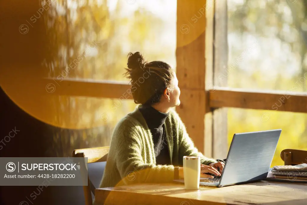 Woman working at laptop looking out window in sunny cafe