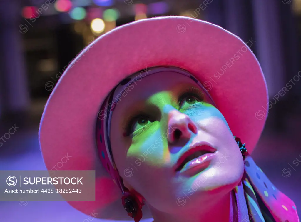 Curious stylish woman in pink hat looking up in neon light