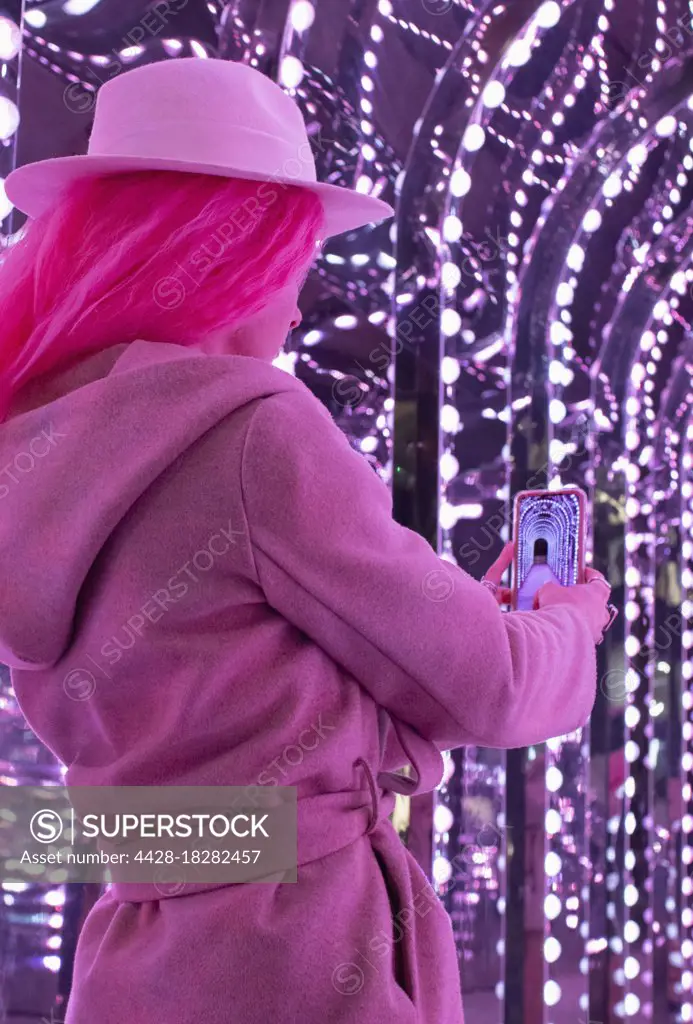Fashionable woman in pink coat using camera phone under lights