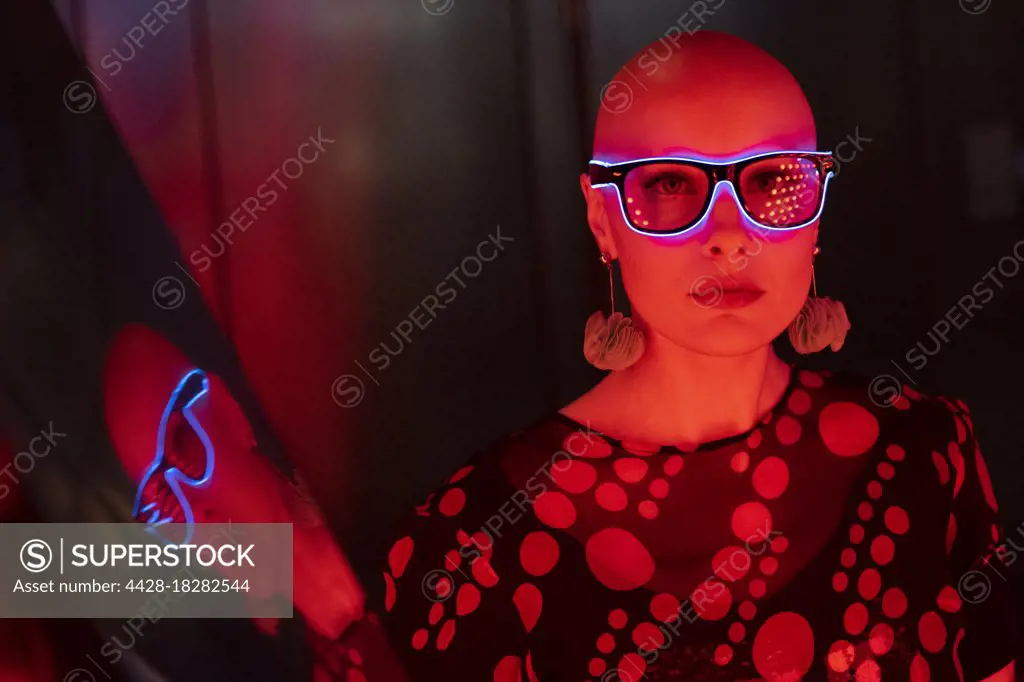 Portrait cool woman with shaved head and neon glasses in red light