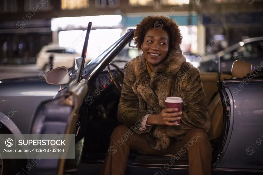 Happy young woman with coffee at convertible in city at night