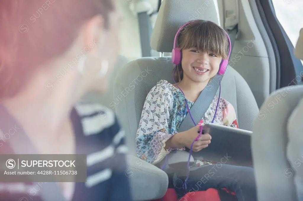 Mother turning and smiling at daughter with headphones and digital tablet in back seat of car