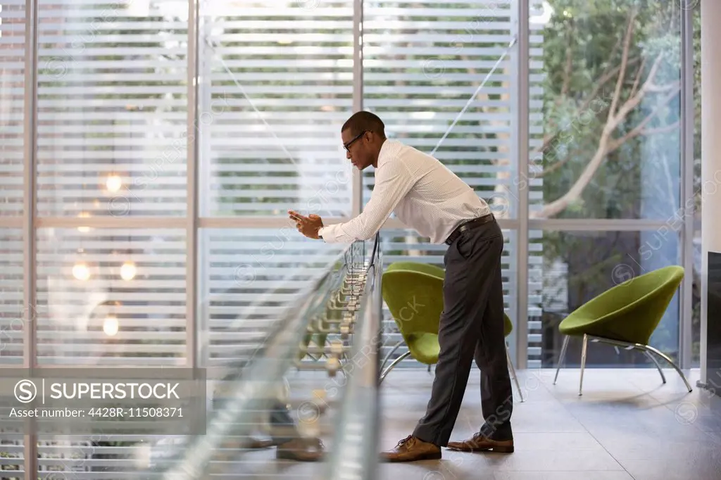 Businessman leaning on railing in office and text messaging with cell phone