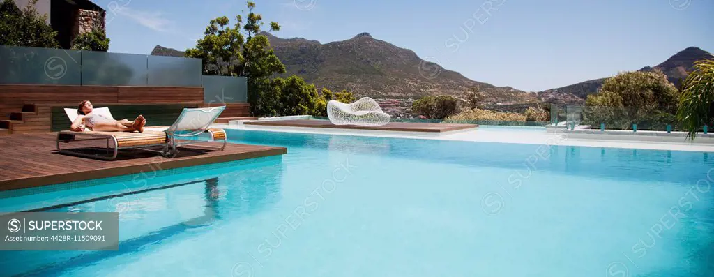 Woman sunbathing on lounge chair next to luxury swimming pool with mountain view