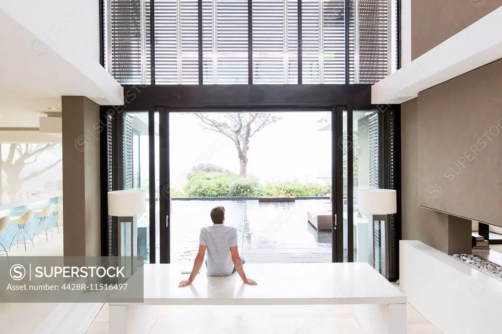 Rear view of man sitting on table and looking at swimming pool through open patio door
