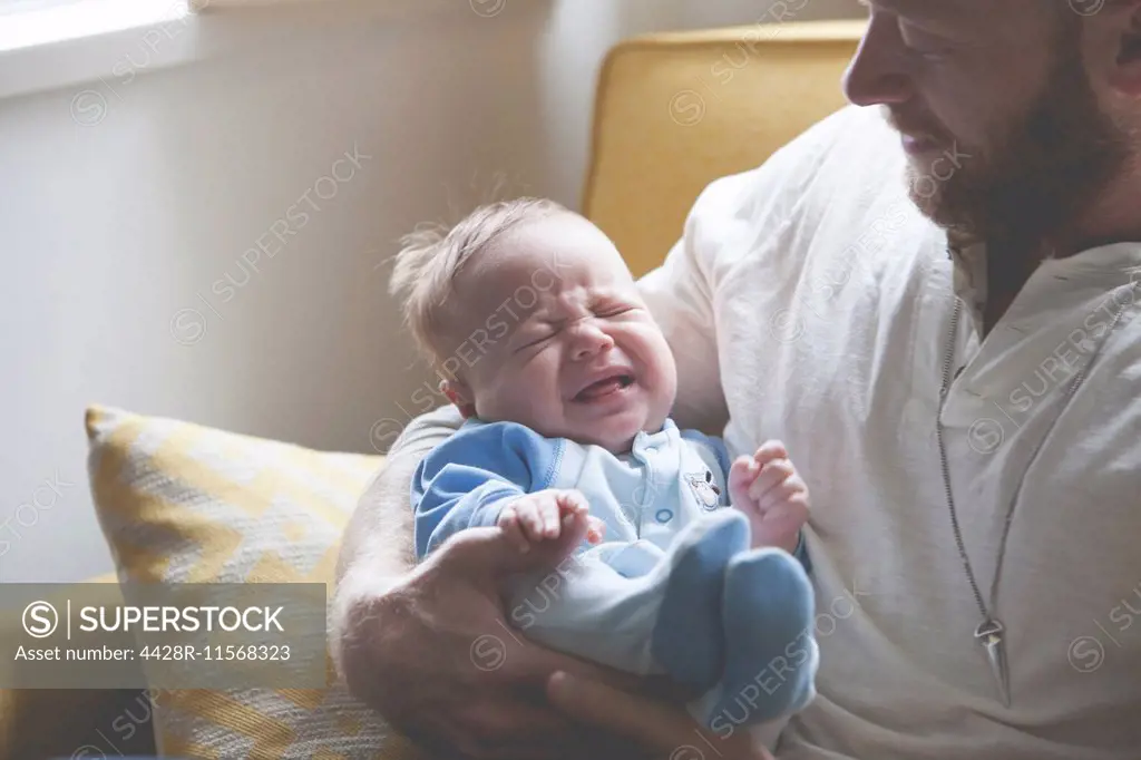 Father holding and looking at crying baby, sitting on sofa