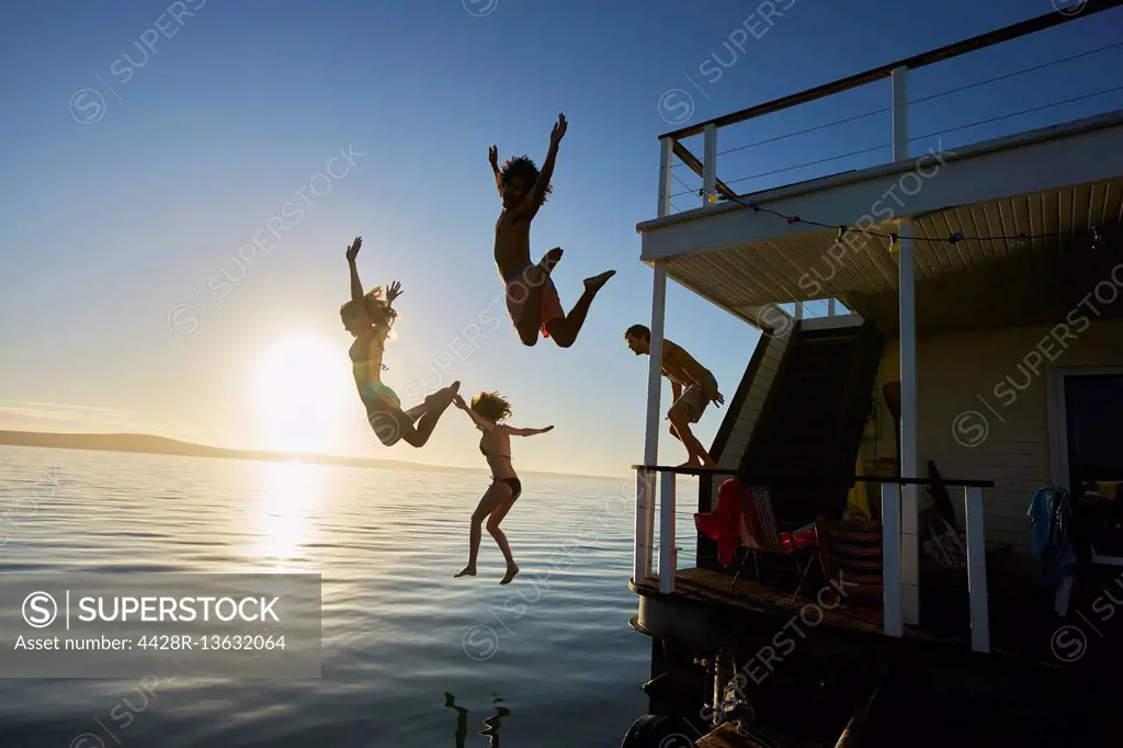 Young adult friends jumping off summer houseboat into sunset ocean