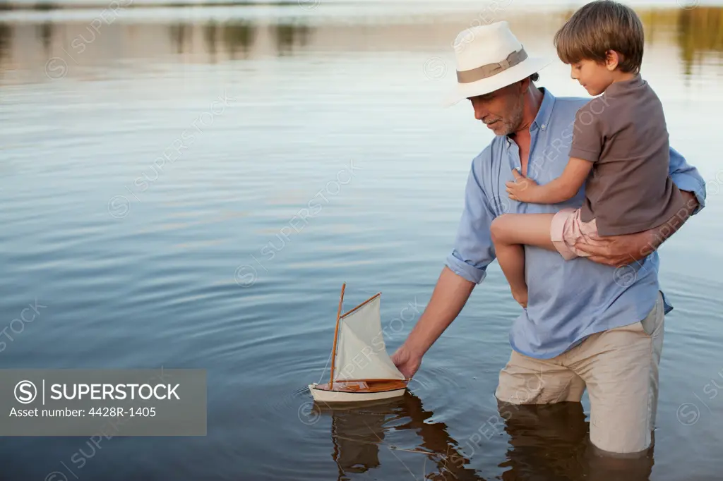 Cape Town, Grandfather and grandson wading in lake with toy sailboat