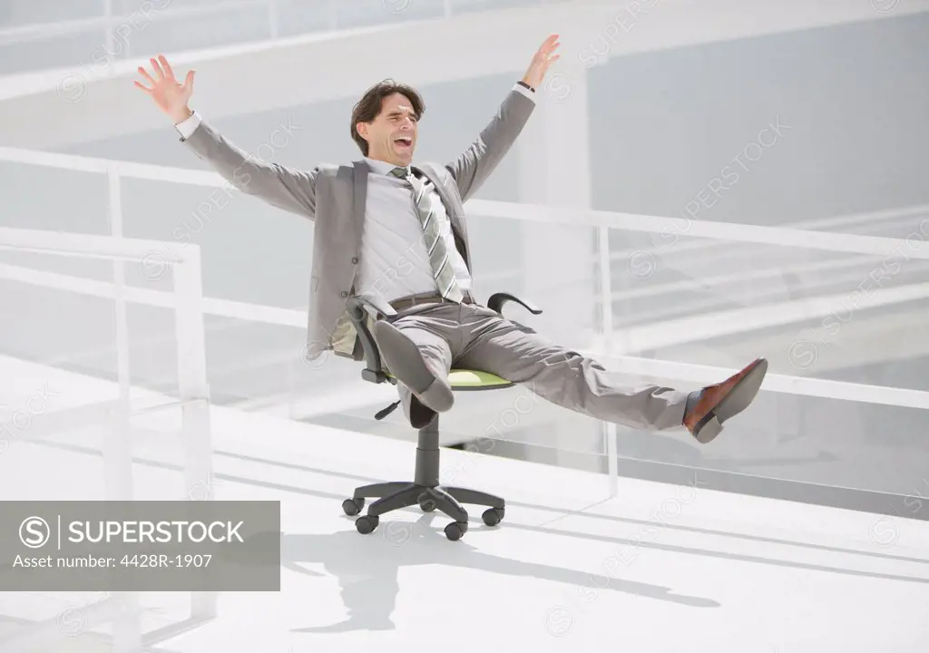 Spain, Carefree businessman sliding down walkway on office chair with wheels
