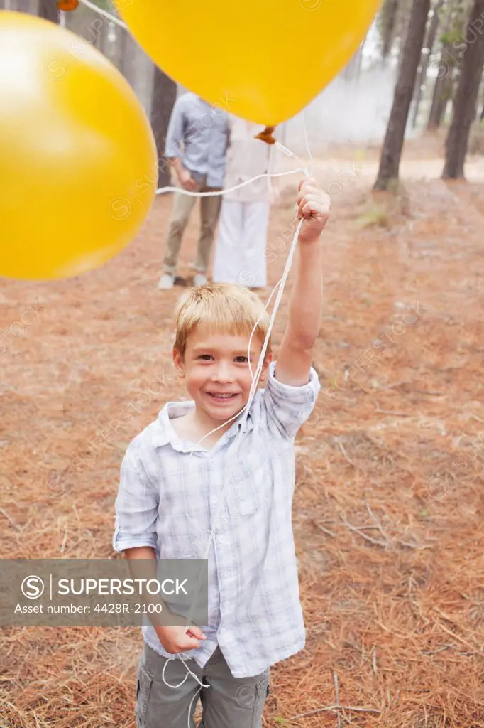 Cape Town, South Africa, Portrait of smiling boy holding balloons in woods with parents in background