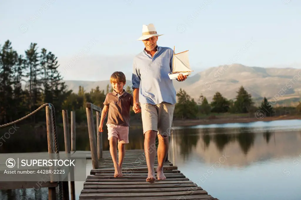 Cape Town, Smiling grandfather and grandson with toy sailboat holding hands and walking along dock over lake