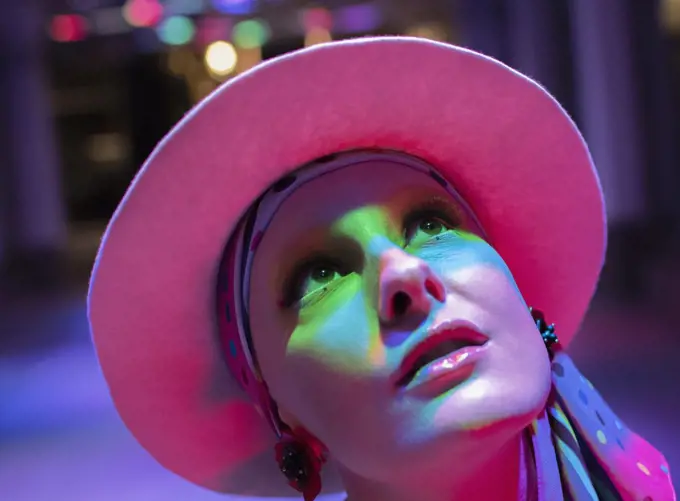 Curious stylish woman in pink hat looking up in neon light