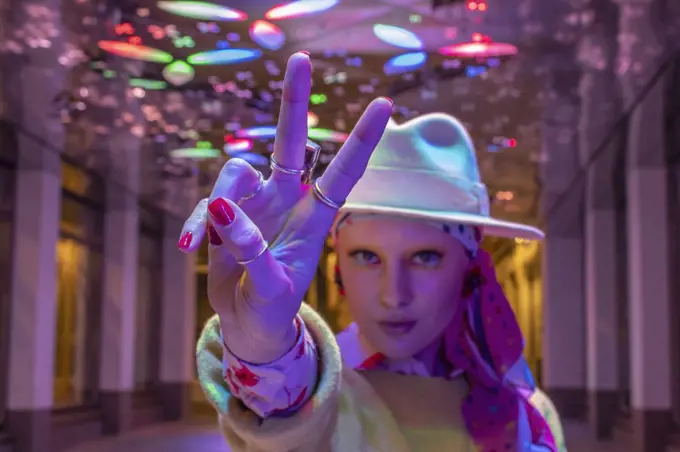 Portrait cool stylish woman in fedora gesturing peace sign under neon
