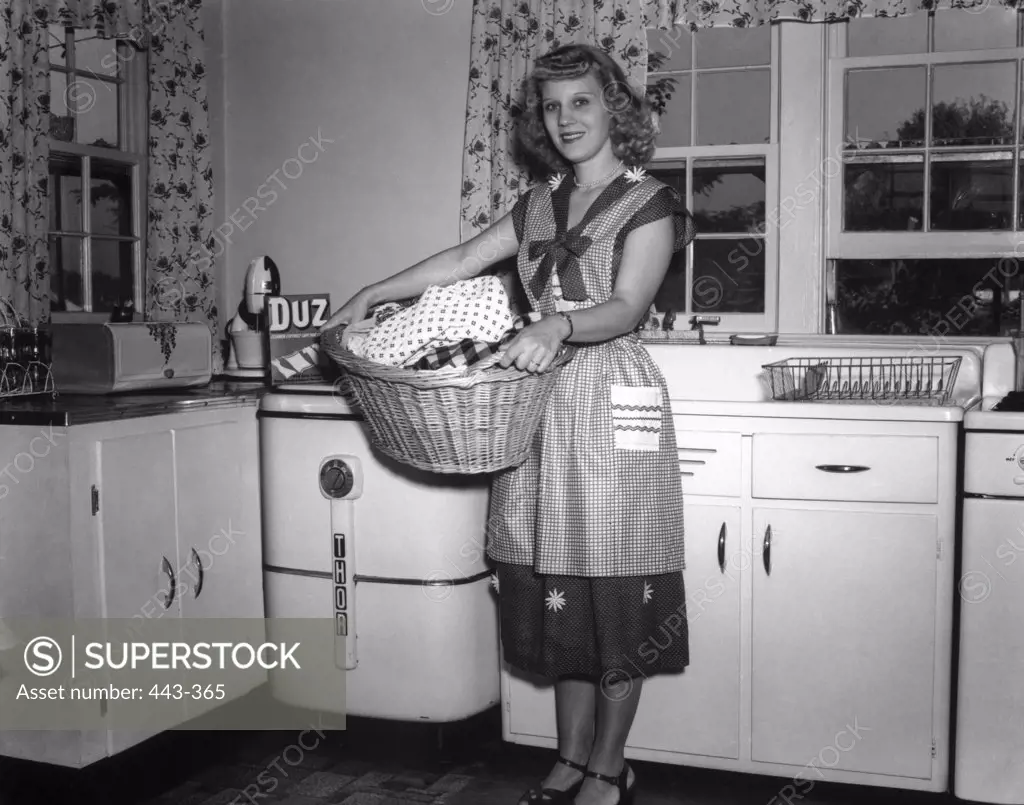 Portrait of a young woman holding a basket full of clothes in a kitchen, 1951