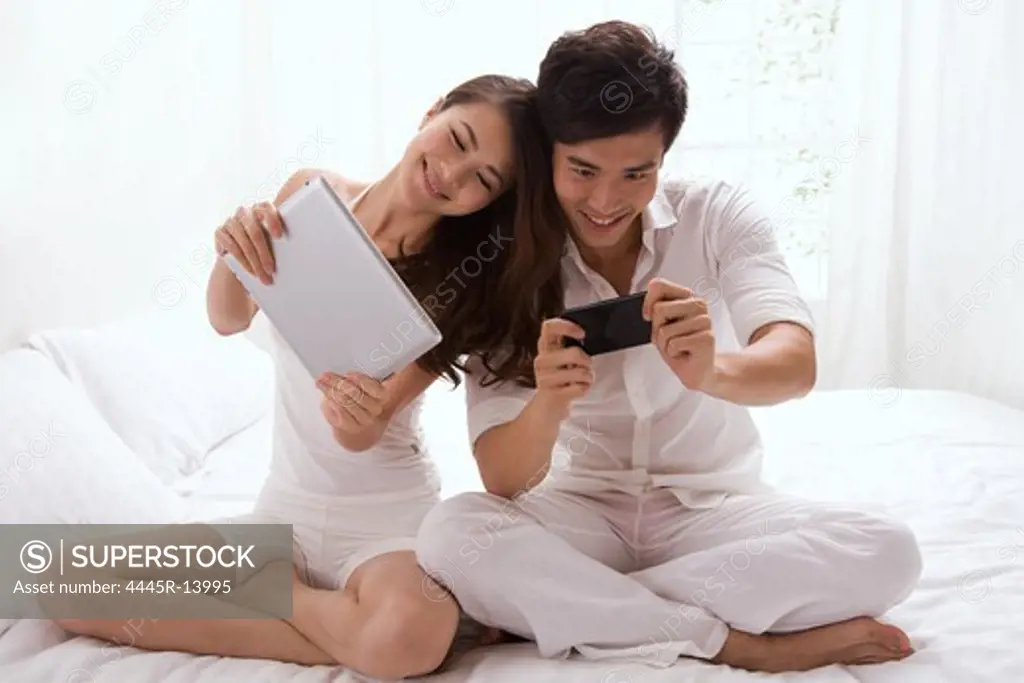 Young couple watching cell phone and tablet computer on bed