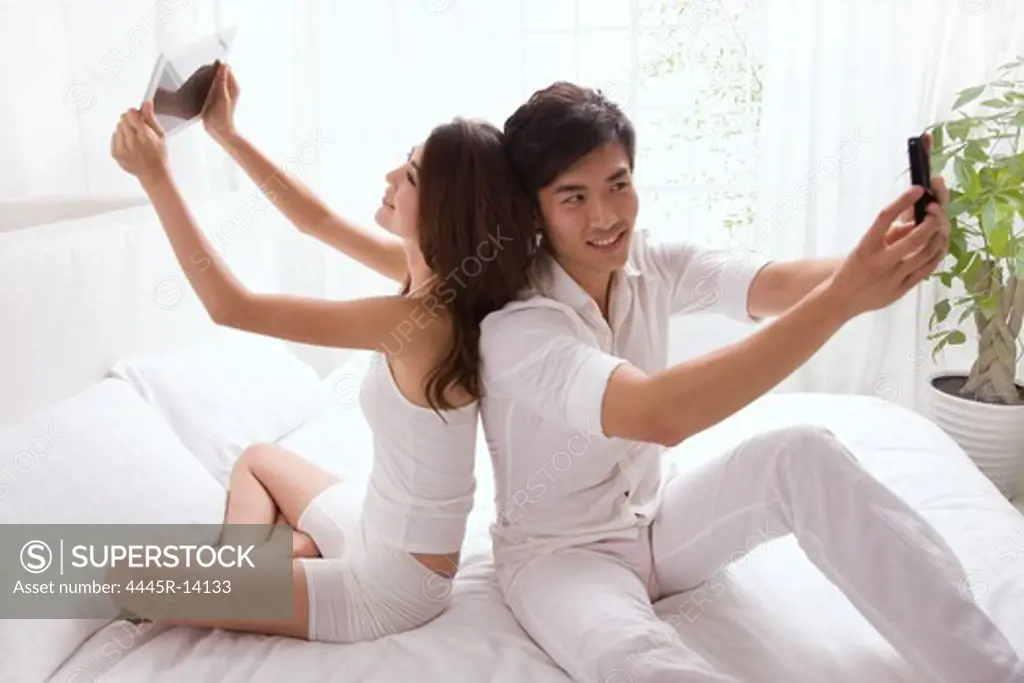 Young couple holding cell phone on bed back to back