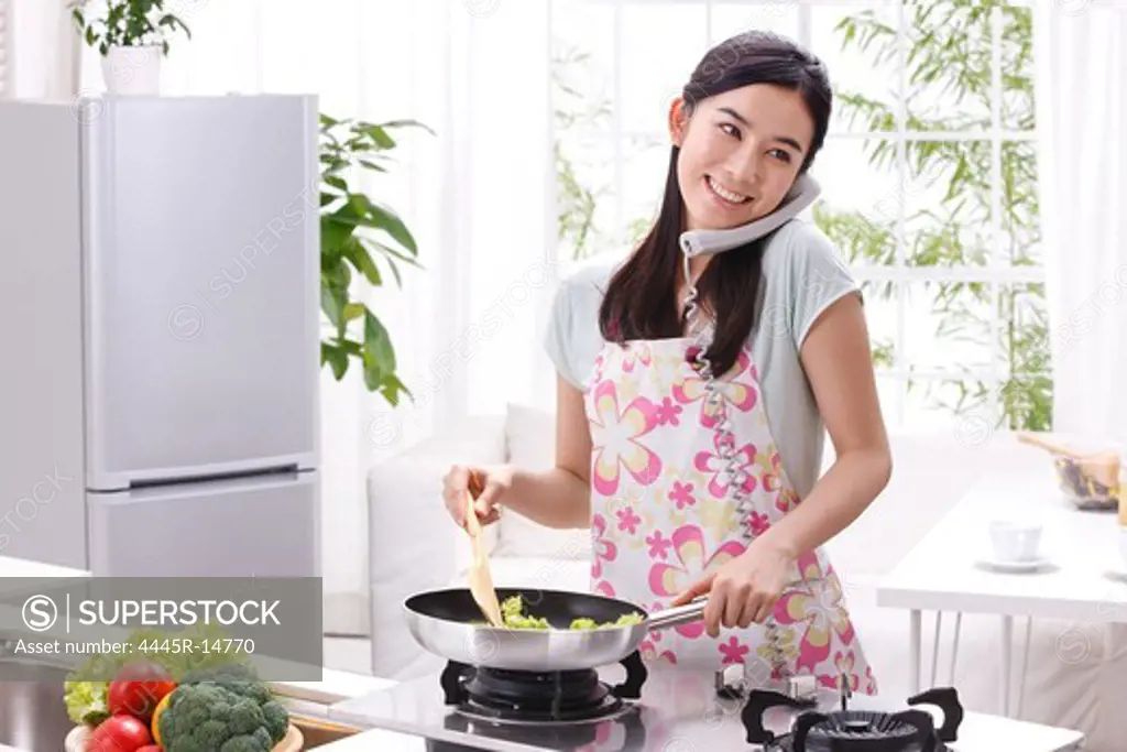 Young woman making phone call and cooking in kitchen