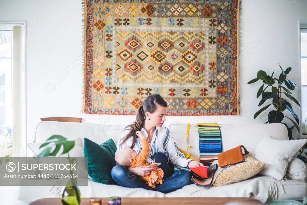 Fashion designer breastfeeding daughter while looking at fabric swatch on sofa in living room