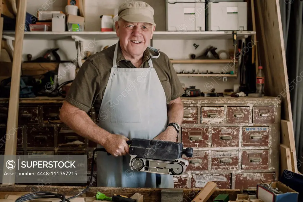 Portrait of confident smiling senior male owner holding power tool at workshop