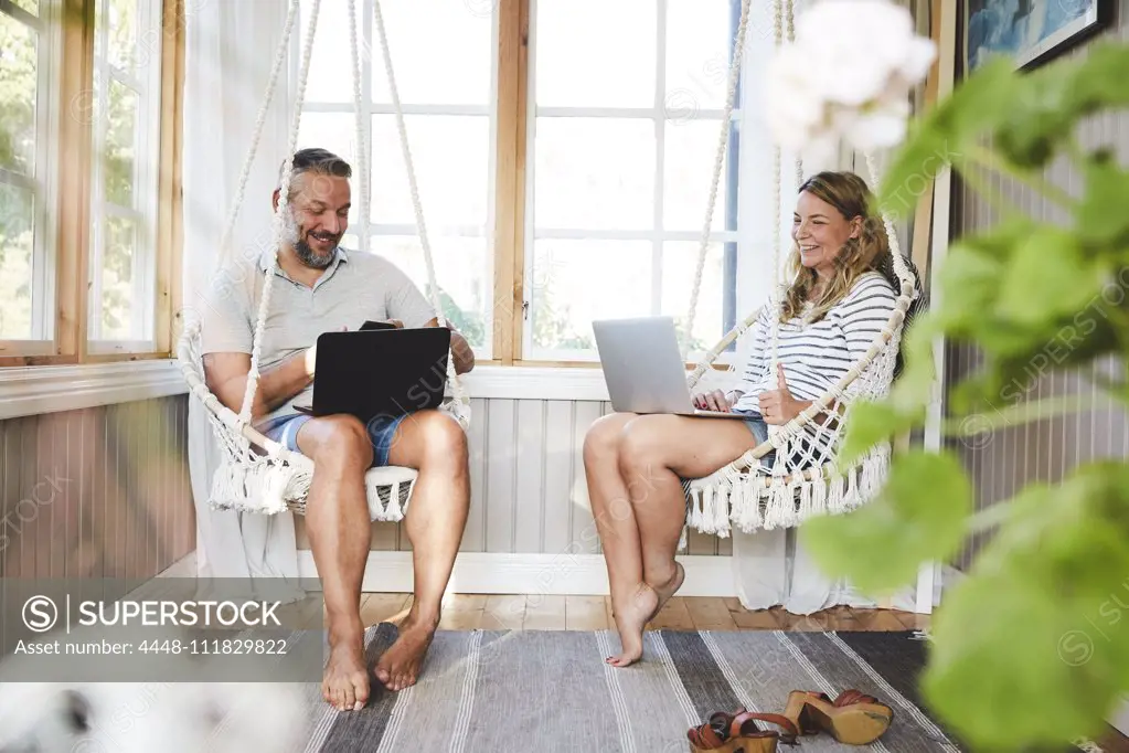 Smiling couple using laptops while sitting on rope swing in log cabin