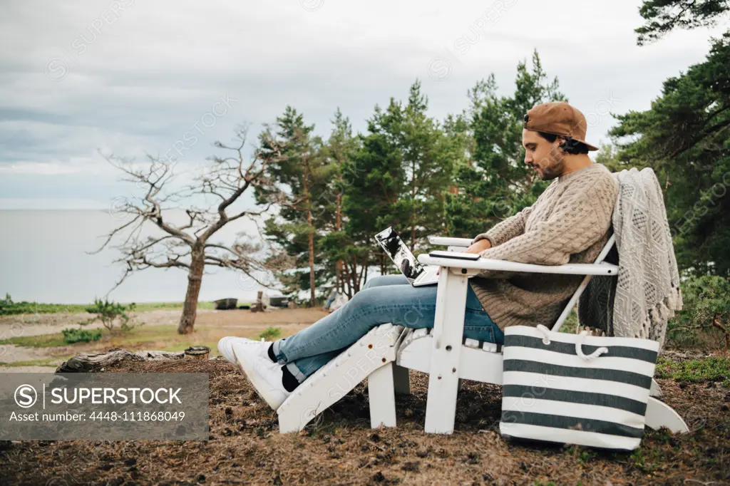 Side view of man using laptop while sitting on Adirondack chair at forest