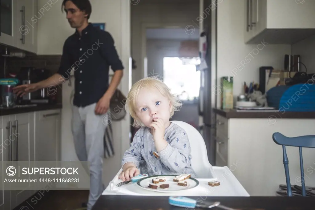 Portrait of blond baby boy eating while sitting on high chair at kitchen with father in background