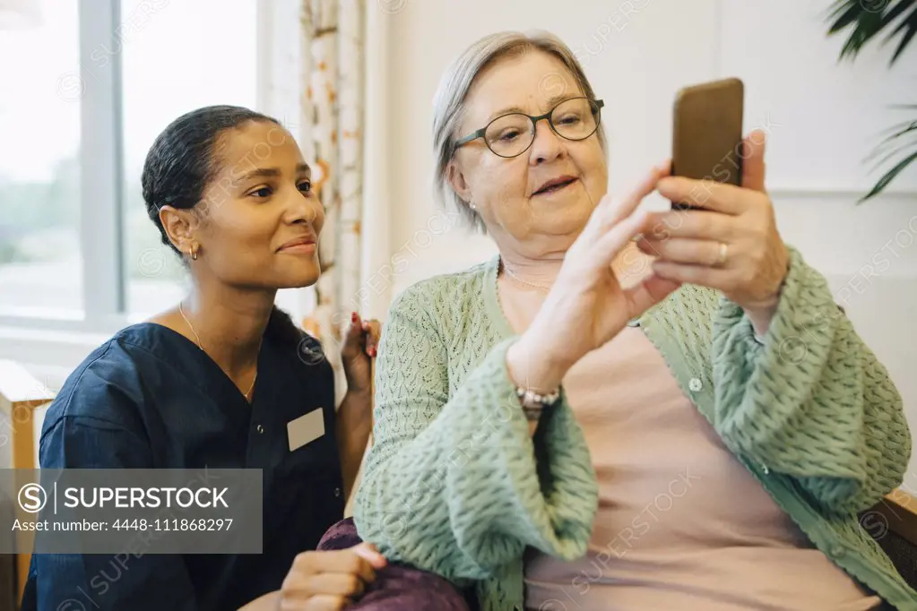 Smiling female caretaker looking at senior woman using mobile phone in nursing home