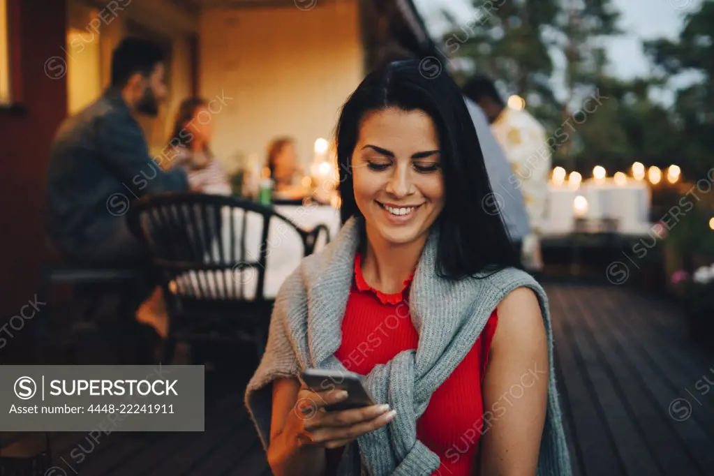Smiling mid adult woman using mobile phone while friends in background during dinner party