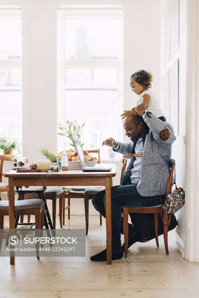 Father carrying daughter on shoulder while using laptop at table in house